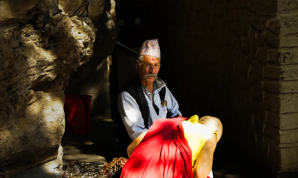 An older man in a Nepali cap and vest sits in a rock niche beside a red cloth, hard sunlight on his face.
