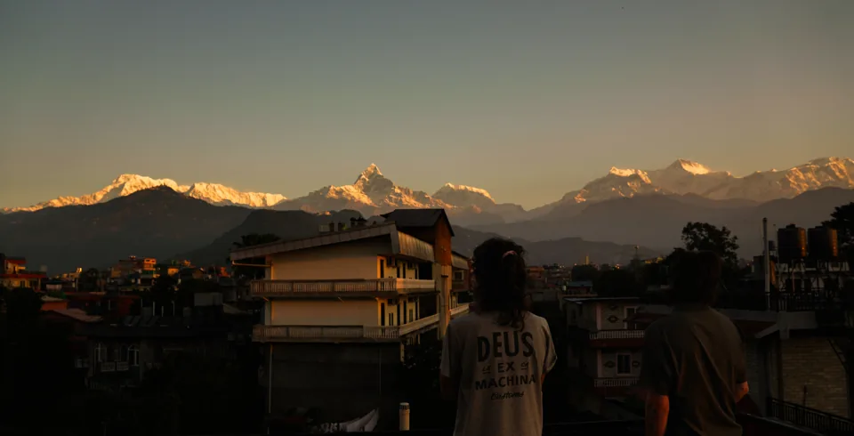 Two people seen from behind on a rooftop at dawn, a snow-covered mountain range along the horizon.