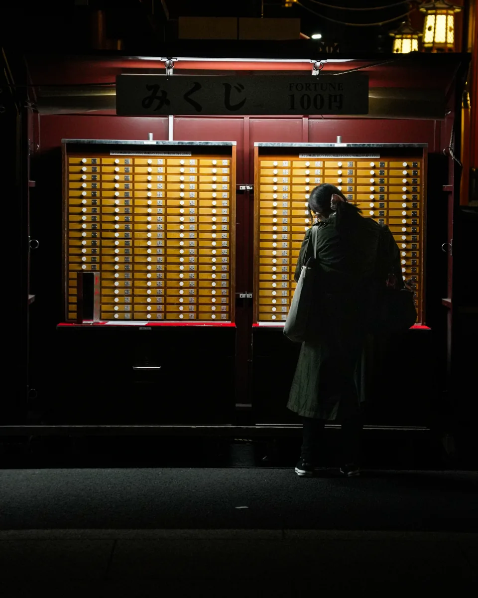 A woman in a green coat stands before red cabinets of small numbered wooden drawers at a fortune stall.
