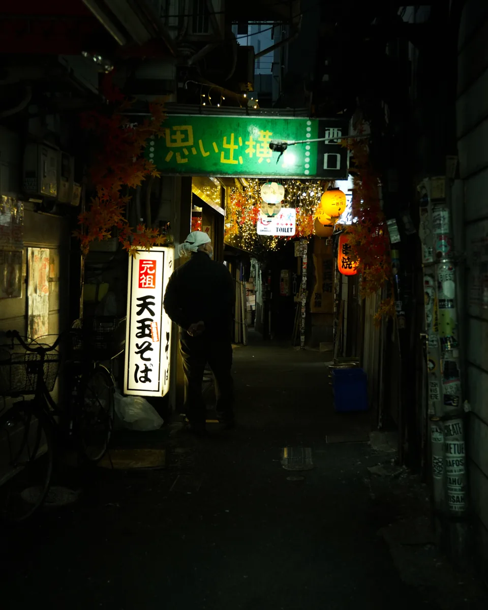 A narrow alley at night lit by lanterns and a green sign, a person walking away past a bicycle.
