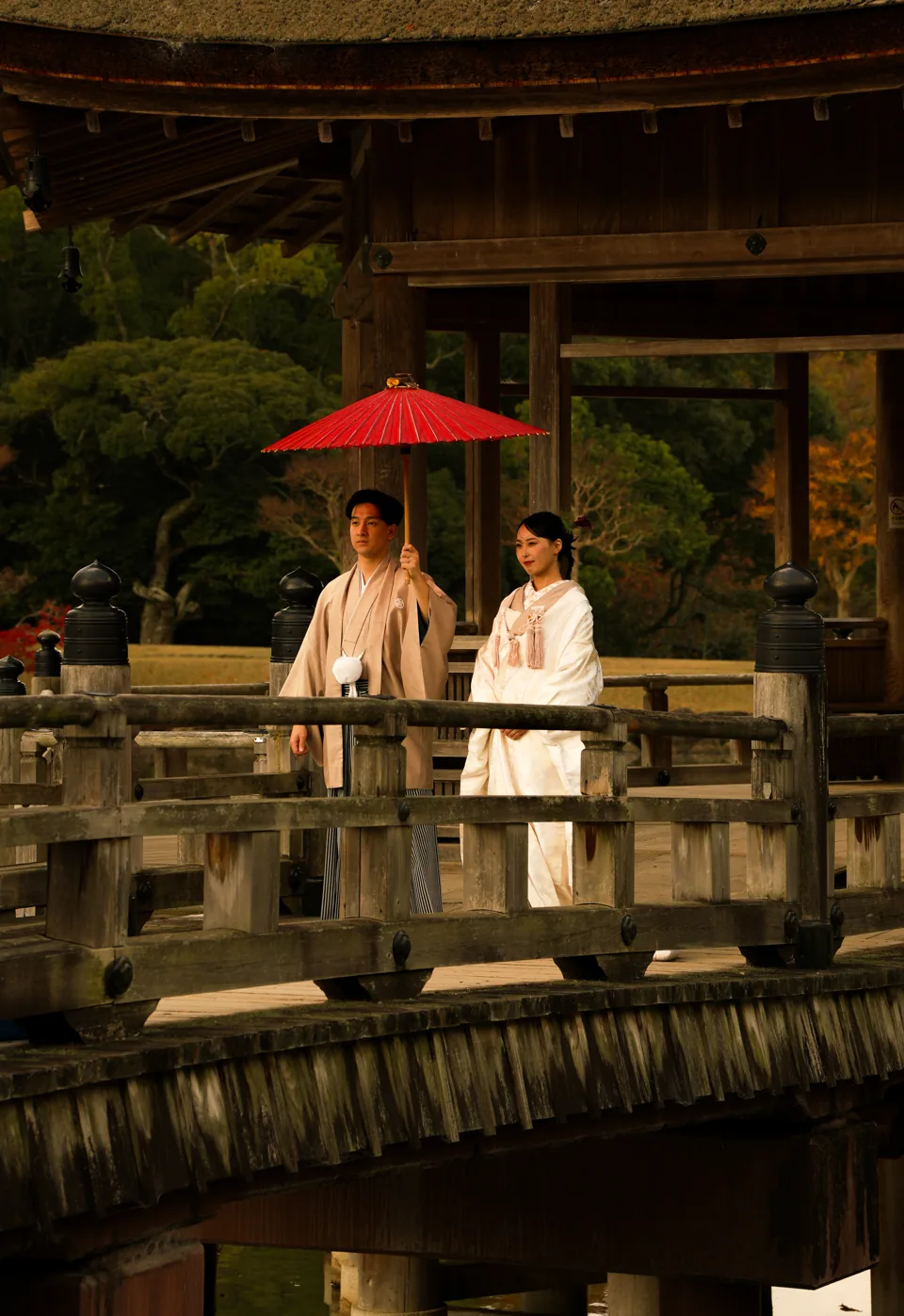 A couple in traditional Japanese wedding dress stand on a wooden bridge under a red paper parasol, a pavilion and autumn trees behind them.