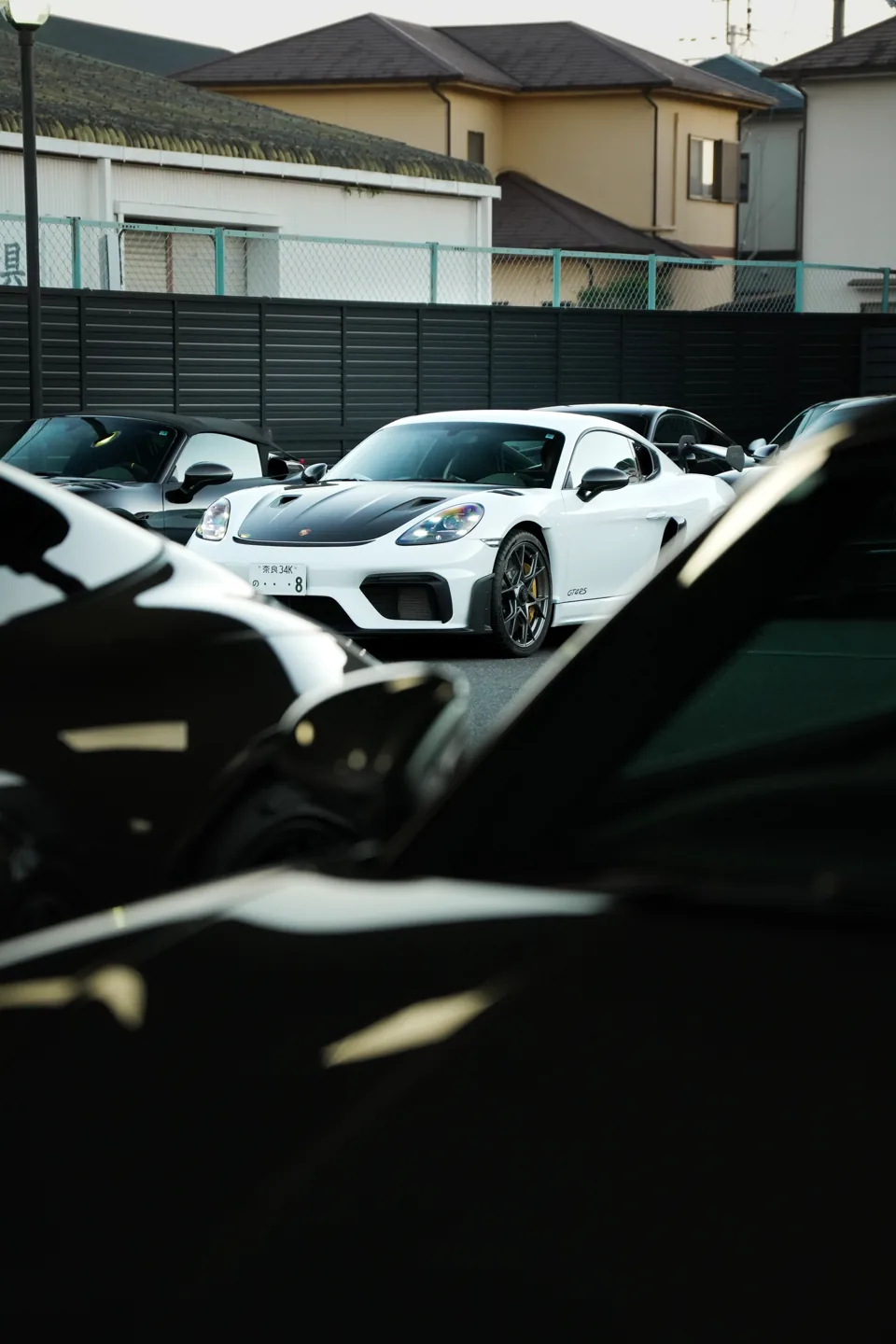 A white Porsche GT4 RS sits among parked cars at dusk, seen through the dark shapes of other cars in the foreground.