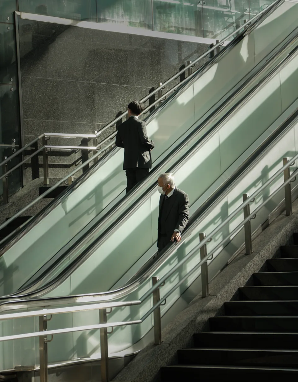 Two men in suits on a glass-sided escalator, one rising, one descending, cold light.