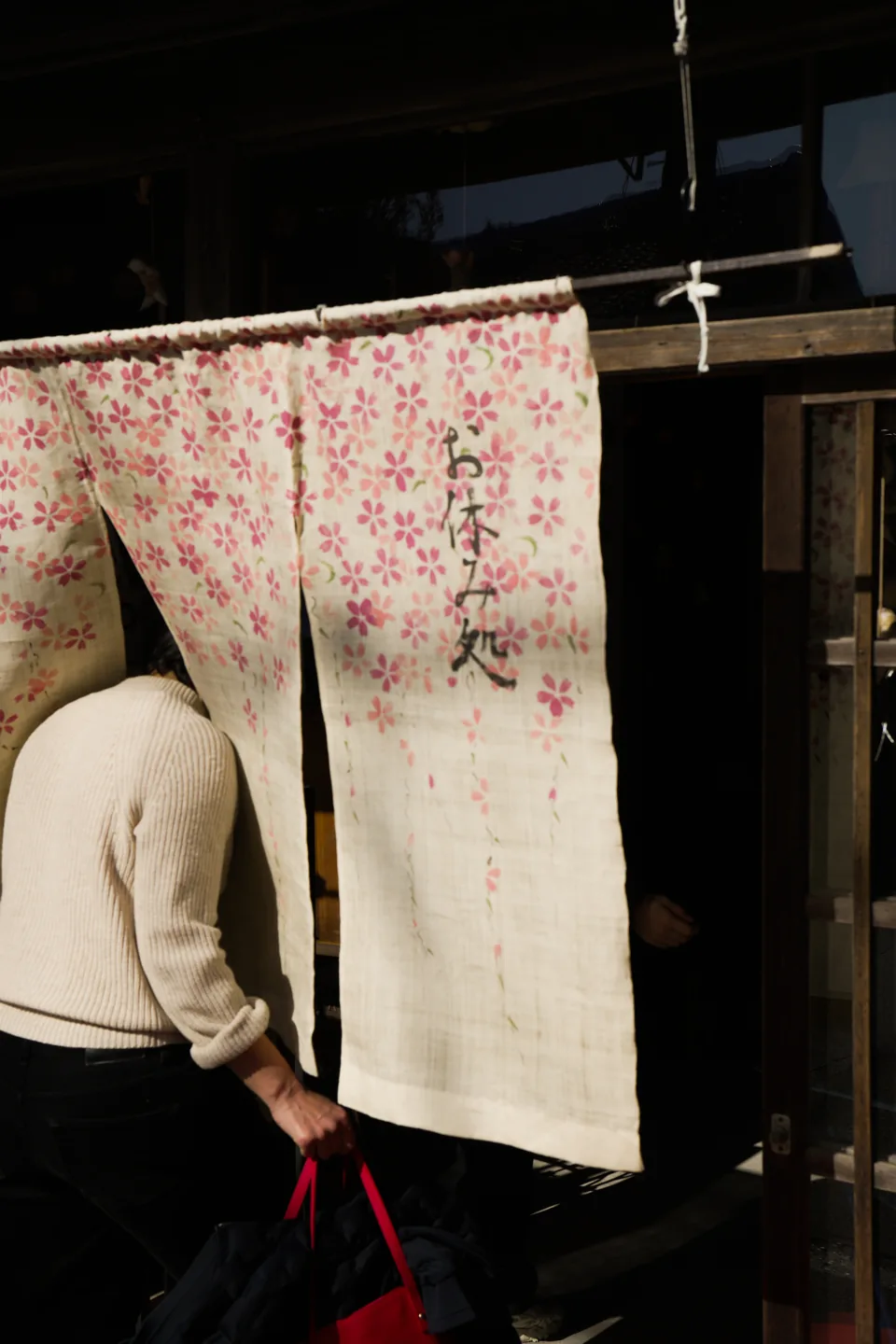 A person in a white sweater passes through a cloth curtain printed with cherry blossoms at a shop entrance.