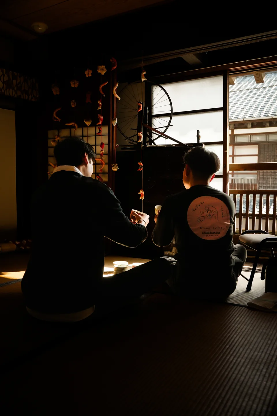 Two men sit on tatami with their backs to the camera, facing a window, tea things on a low table.