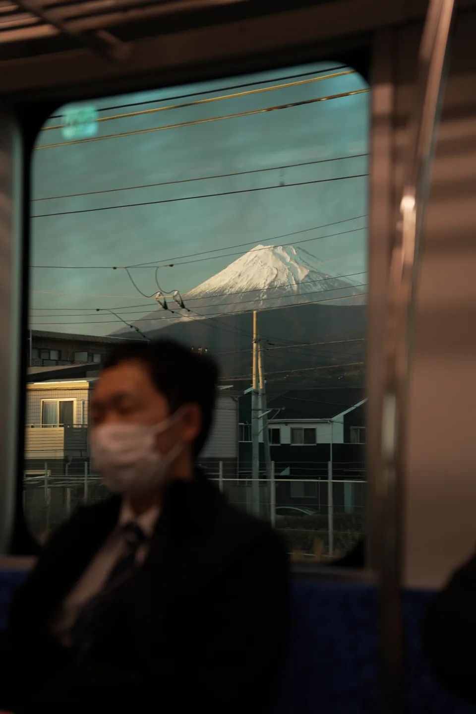 Snow-capped Mount Fuji seen through a train window, power lines crossing the glass, a masked passenger blurred in the foreground.