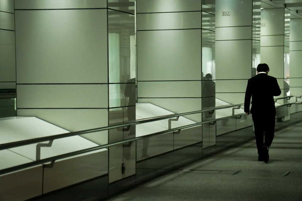 A man in a dark suit walks away down a mirrored corporate corridor, gate numbers on the glass panels beside him.