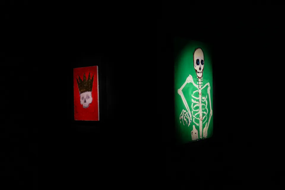 Two skull paintings glow under spotlights in a dark gallery room, part of a Johnny Depp art exhibition - a crowned skull on red, a full skeleton on green.