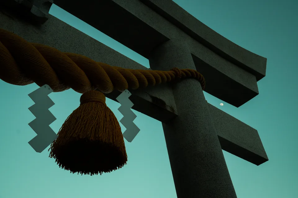 Close view of a torii gate's thick straw shimenawa rope with zigzag paper streamers, a faint moon low in a teal sky beside the crossbeam.