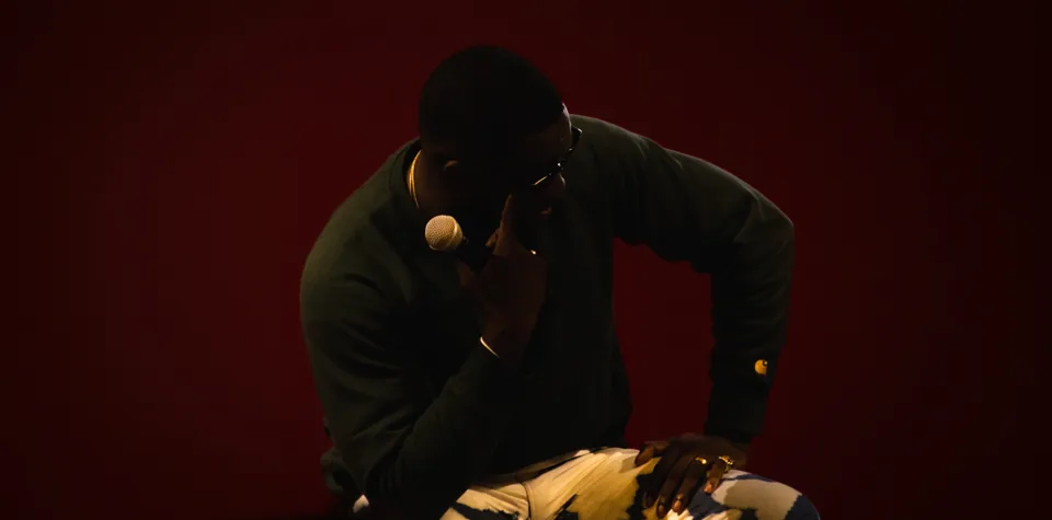 A singer seated on stage against a dark red wall, head bowed, a microphone in one hand, gold rings catching the light.