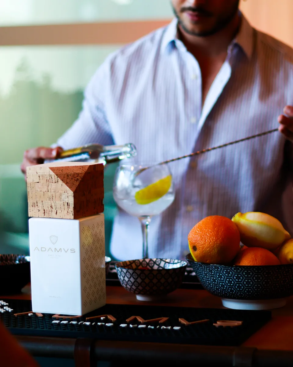 A man in a striped shirt pours gin into a glass at a bar beside a white bottle box and a bowl of citrus.