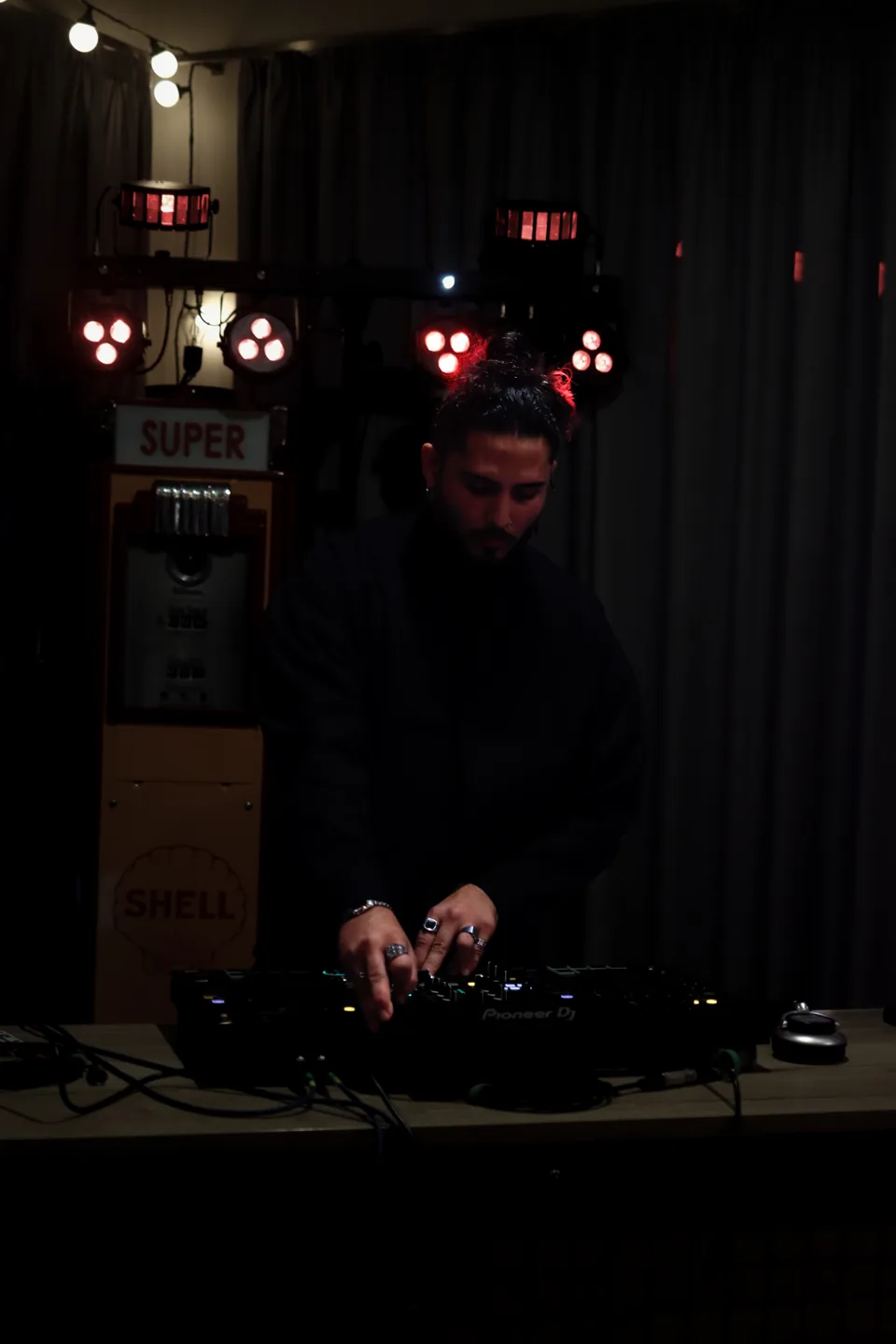 A DJ bends over a mixer under red lights, an illuminated sign behind him.