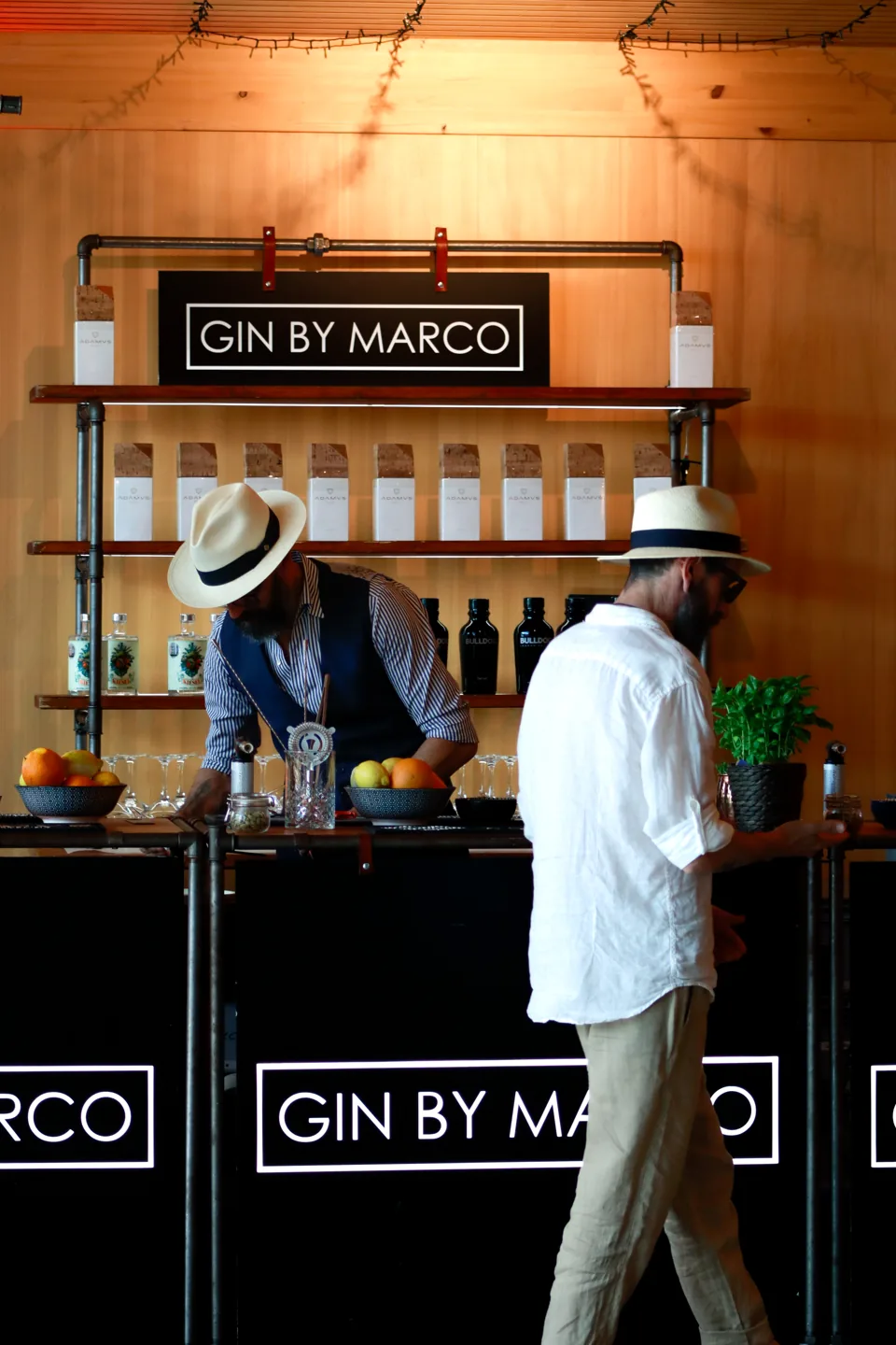 Two men in panama hats behind a bar stand, bottles on shelves, a sign above.