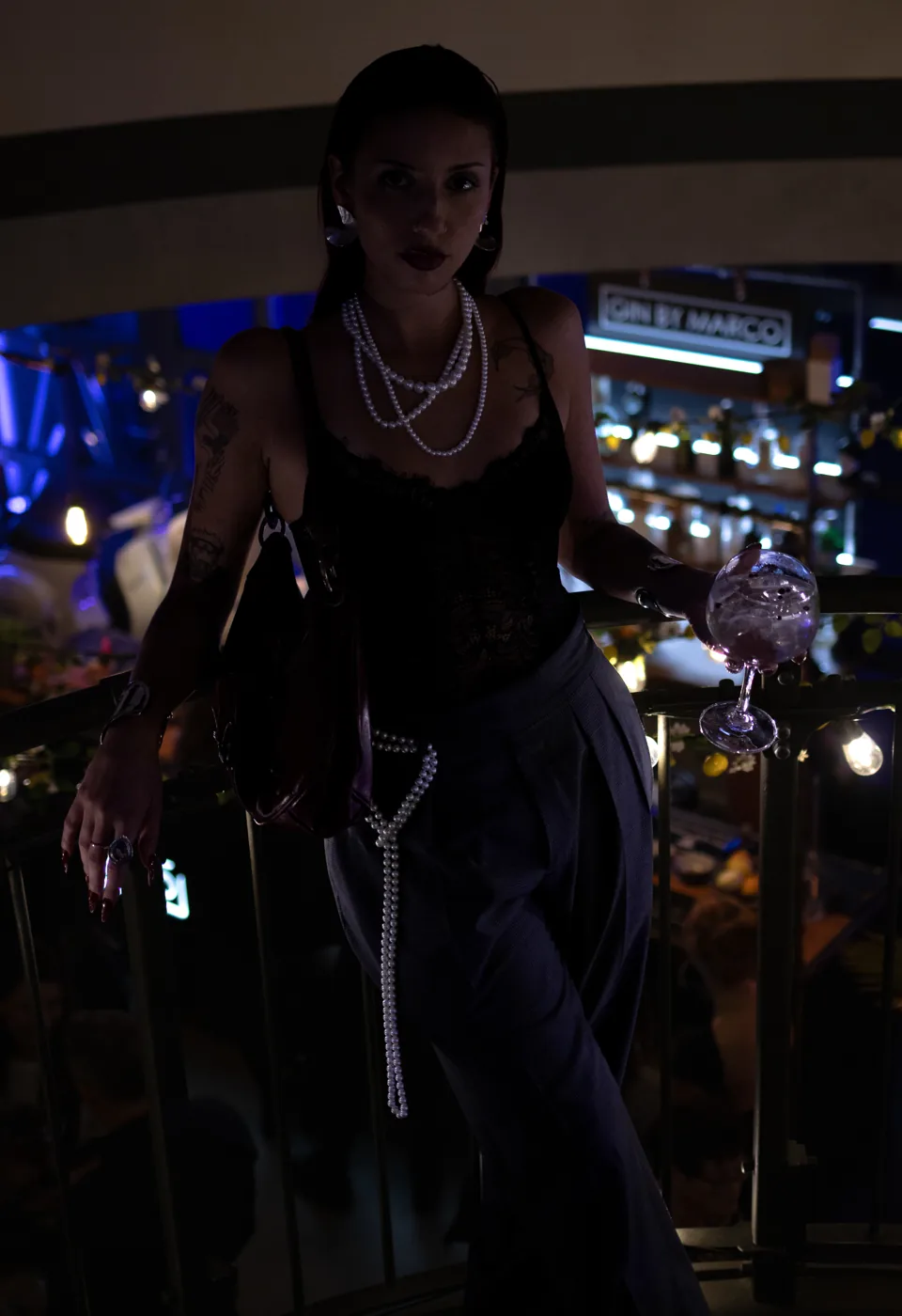 A woman in a black lace top and pearls holds a glass at a bar counter at night.