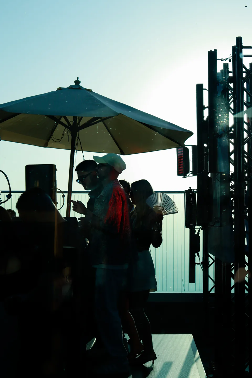 People on a rooftop at sunset under parasols, a sound rig against the light.