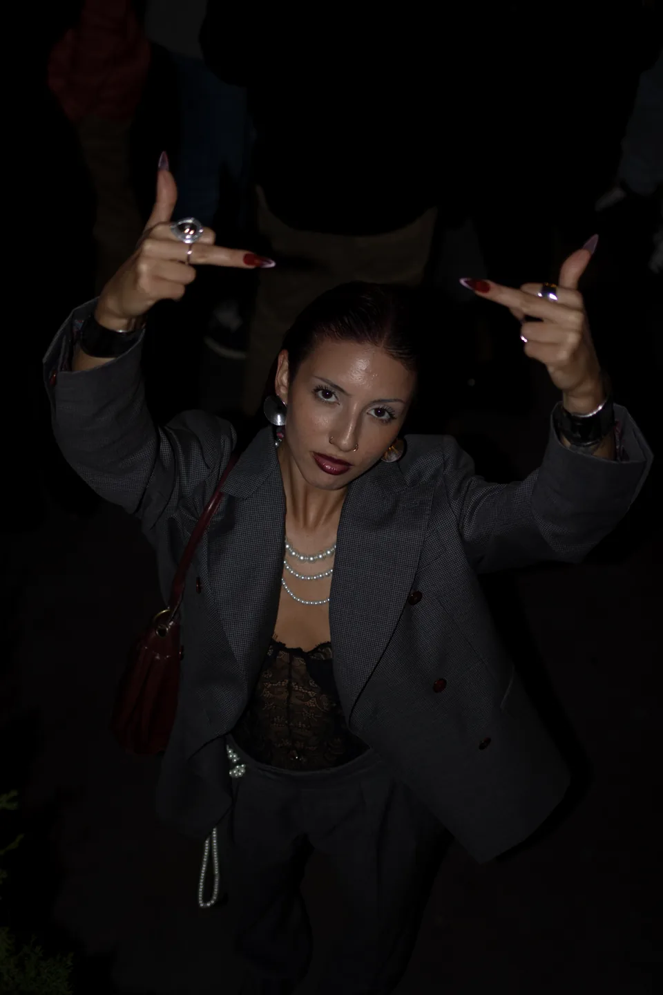 A woman in a grey blazer and pearls raises both hands toward the camera at night, a red bag on her shoulder.
