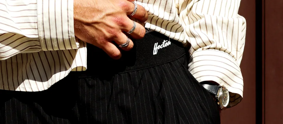 Close view of a hand resting on a black belt buckle, pinstriped trousers and a striped shirt cuff, a watch on the wrist.