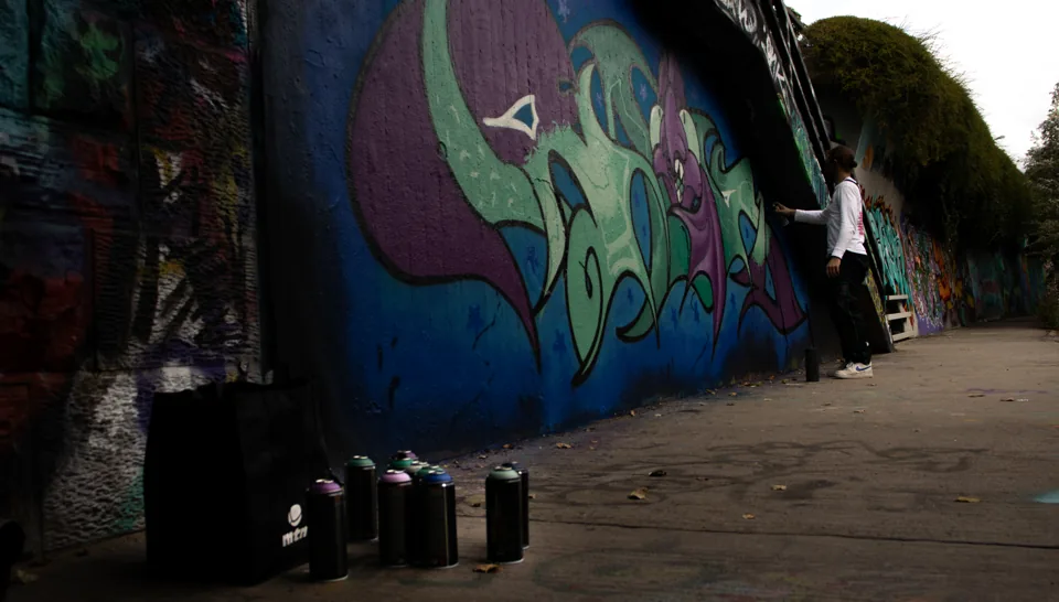 A person in a white long-sleeve top sprays green and purple graffiti lettering on a long wall, spray cans lined up on the pavement, a tree-lined path stretching into the distance.
