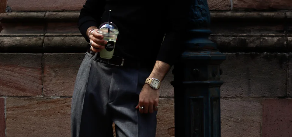 Close view of a hand holding an iced drink with a straw, a watch and rings, a sandstone wall behind.