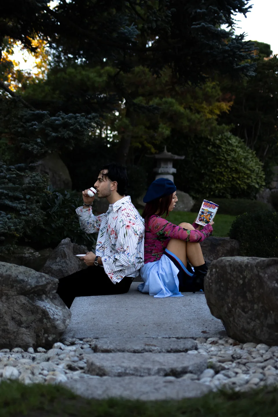 A man and a woman sit back to back on a stone path in a garden; he drinks from a small cup, she reads a comic, both in patterned clothes.