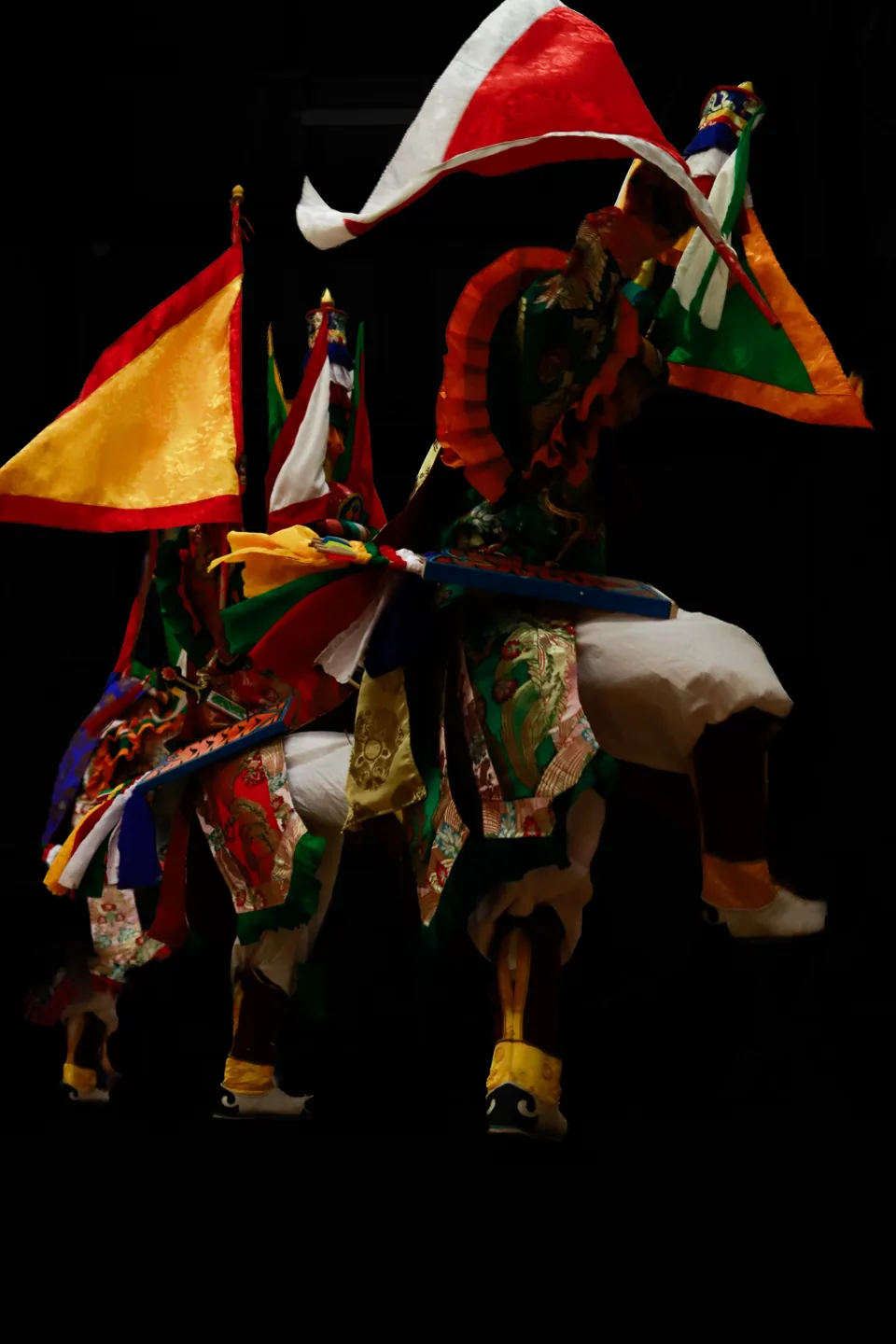 A performer in an embroidered costume with tall flags on the back stands against black.