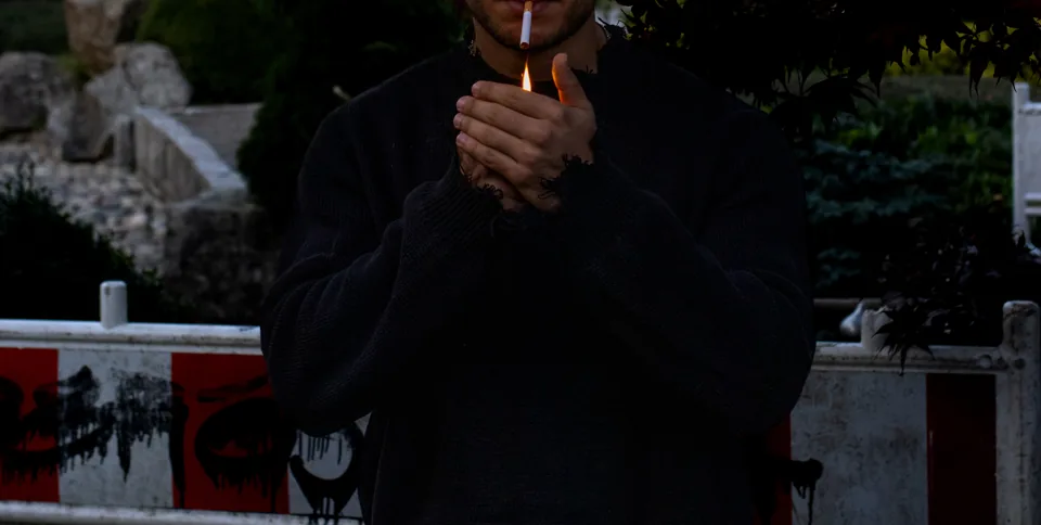 Close view of a person in a black sweater cupping a lit lighter to a cigarette, a striped barrier out of focus behind.