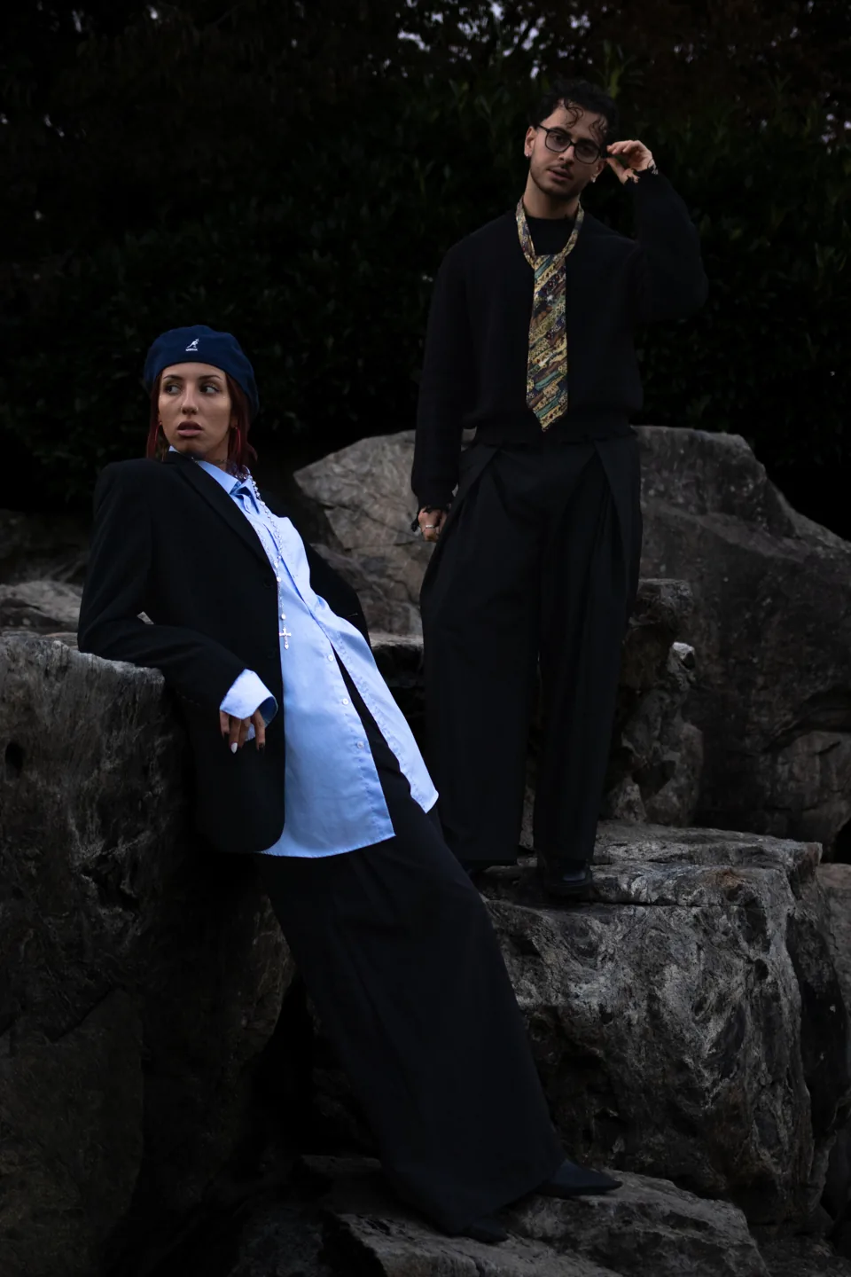 A woman in an oversized black blazer and blue shirt leans against a rock while a man in a black suit and patterned tie stands behind her among boulders.
