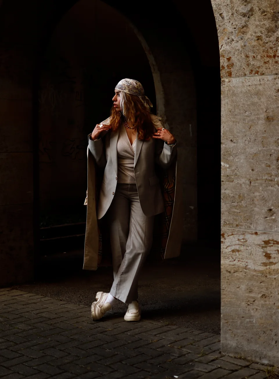 A woman in a beige suit and headscarf stands beneath a stone archway, looking to one side, white sneakers on cobblestones.
