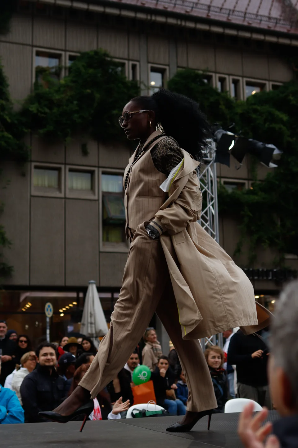A model in a beige trench coat mid-stride on an outdoor runway, photographers and spectators along the edge.