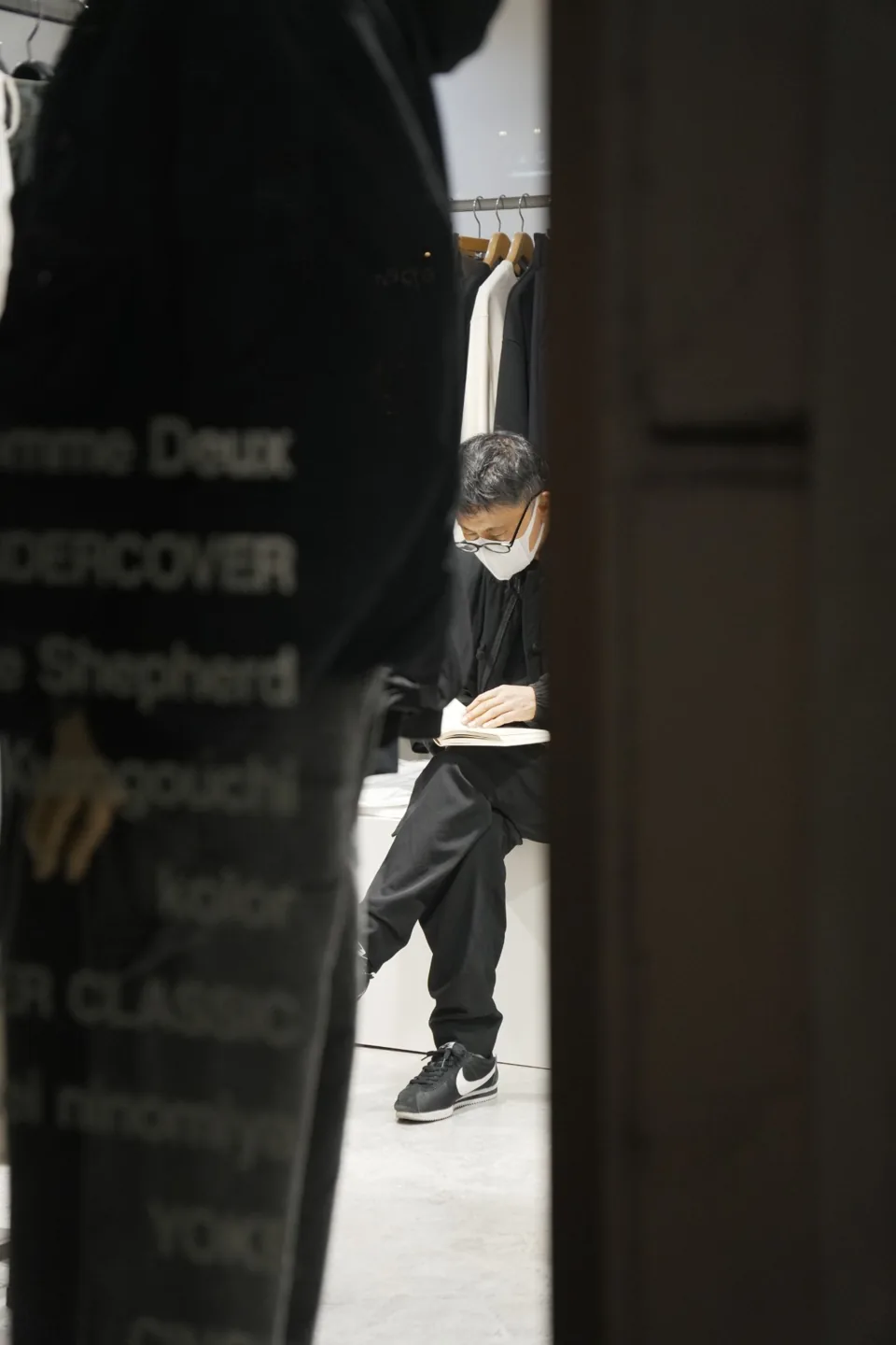 A man in dark clothing sits reading a book in a clothing boutique, masked, one foot on a low platform, racks of black garments behind him.