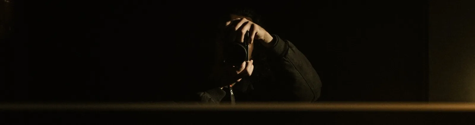 A photographer in a dark room holds his camera to his face, a warm strip of light along the bottom edge.